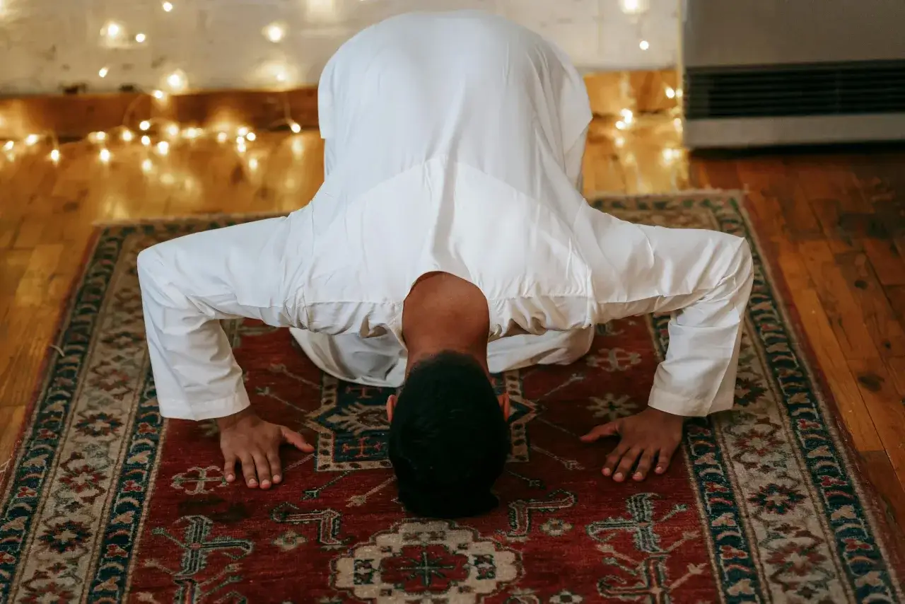 Ein Mann in wei&szlig;er Kleidung beugt sich in **Taqwa** im Islam auf einem Teppich. Lichterketten im Hintergrund schaffen eine friedliche Atmosph&auml;re.