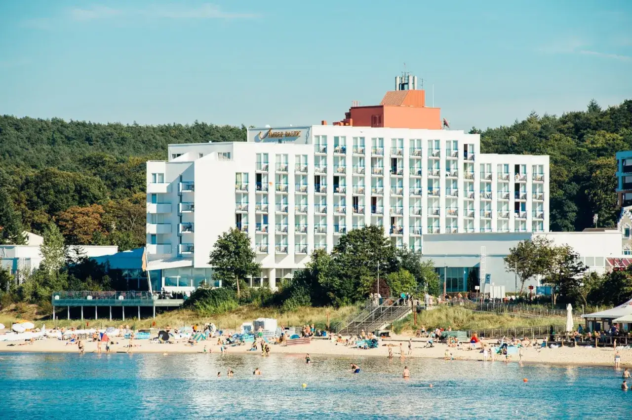 Hotel Amber Baltic Międzyzdroje z widokiem na plażę i morze. Ludzie odpoczywają na piasku i kąpią się w wodzie.