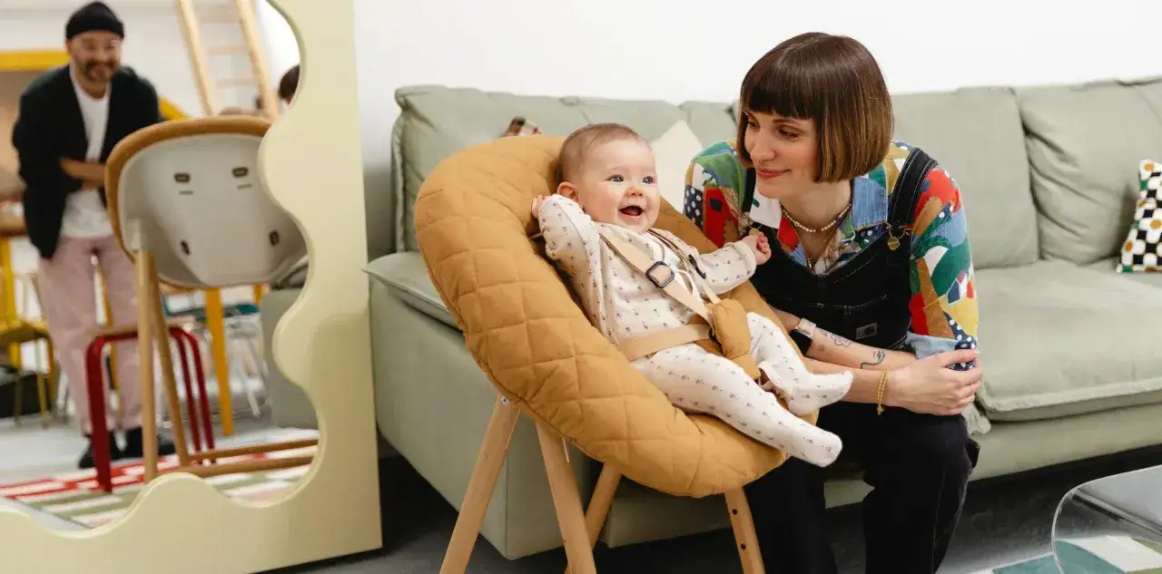 Ein fröhliches Baby von 9 Monaten sitzt in einem gemütlichen Hochstuhl. Eine Frau lächelt das Kind an.