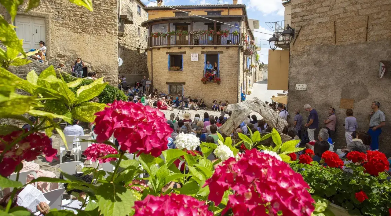 Fiori rosa e bianchi incorniciano una piazza dove la gente si riunisce per un evento. Un uomo e una donna affacciati da una finestra sembrano intrattenere la folla. Un cartello indica 
