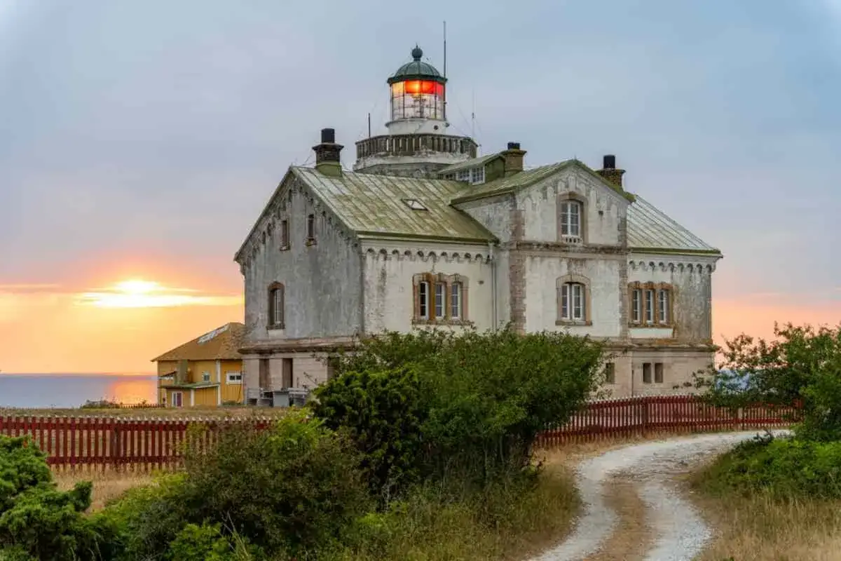 Det gamla fyret på Gotland lyser upp skymningen vid havet. En grusväg leder fram till det vita huset med rött staket.
