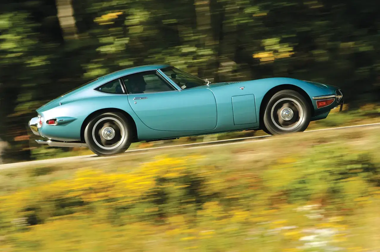 Ein t&uuml;rkisfarbener Toyota 2000GT rast auf einer Stra&szlig;e, umgeben von verschwommenem Gr&uuml;n und gelben Blumen. Der Preis dieses Klassikers ist hoch.