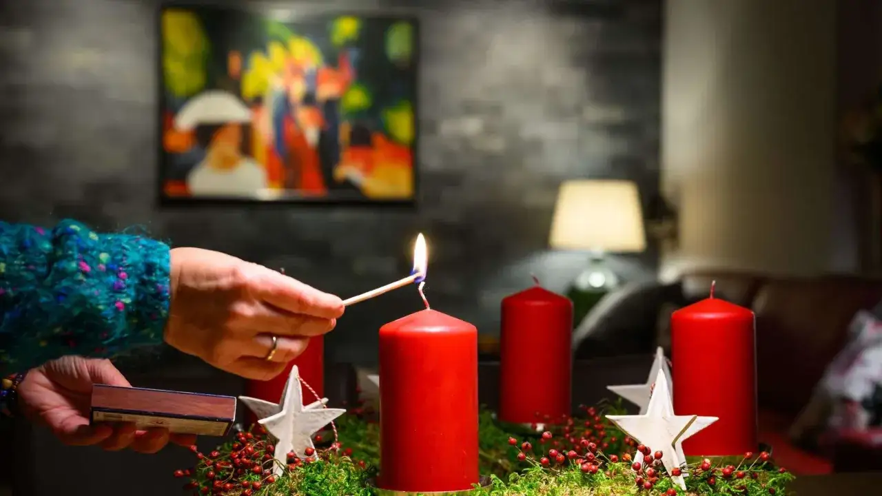 Die dritte Kerze des Adventskranzes wird angez&uuml;ndet. Rote Kerzen, Moos und Sterne schm&uuml;cken den Kranz.