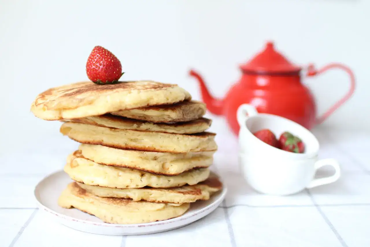Pile de pancakes sans gluten sur une assiette, garnie d'une fraise. Une théière rouge et des fraises dans une tasse sont en arrière-plan.