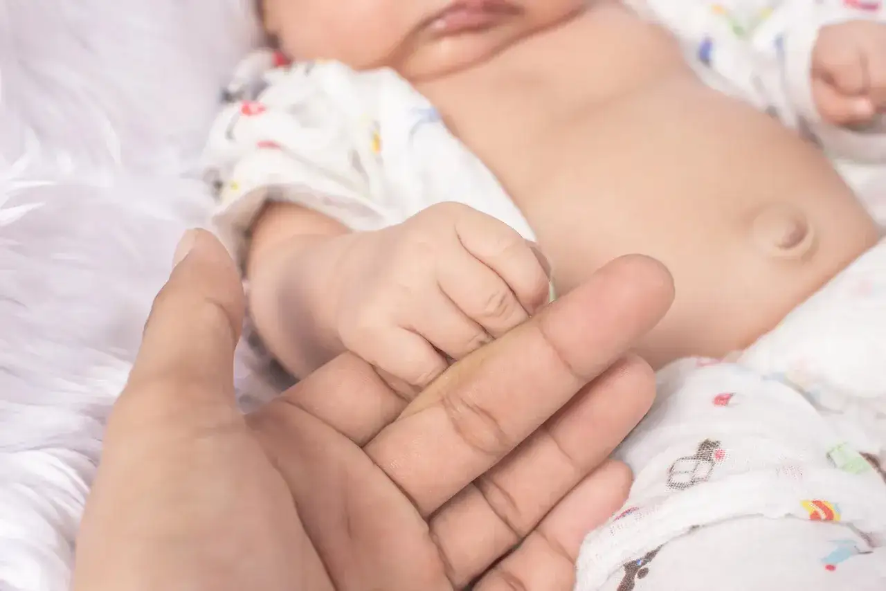 Dłoń rodzica obejmuje maleńką rączkę dziecka, na brzuszku widoczna przepuklina pępkowa u niemowlaka.