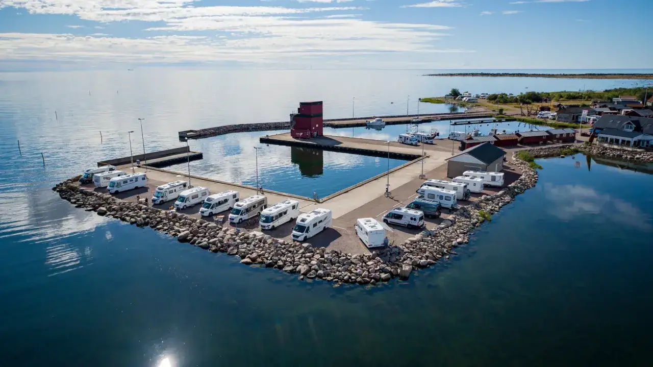Husvagnssemester Sverige: husbilar och husvagnar parkerade vid en hamn med blått vatten och en röd byggnad.