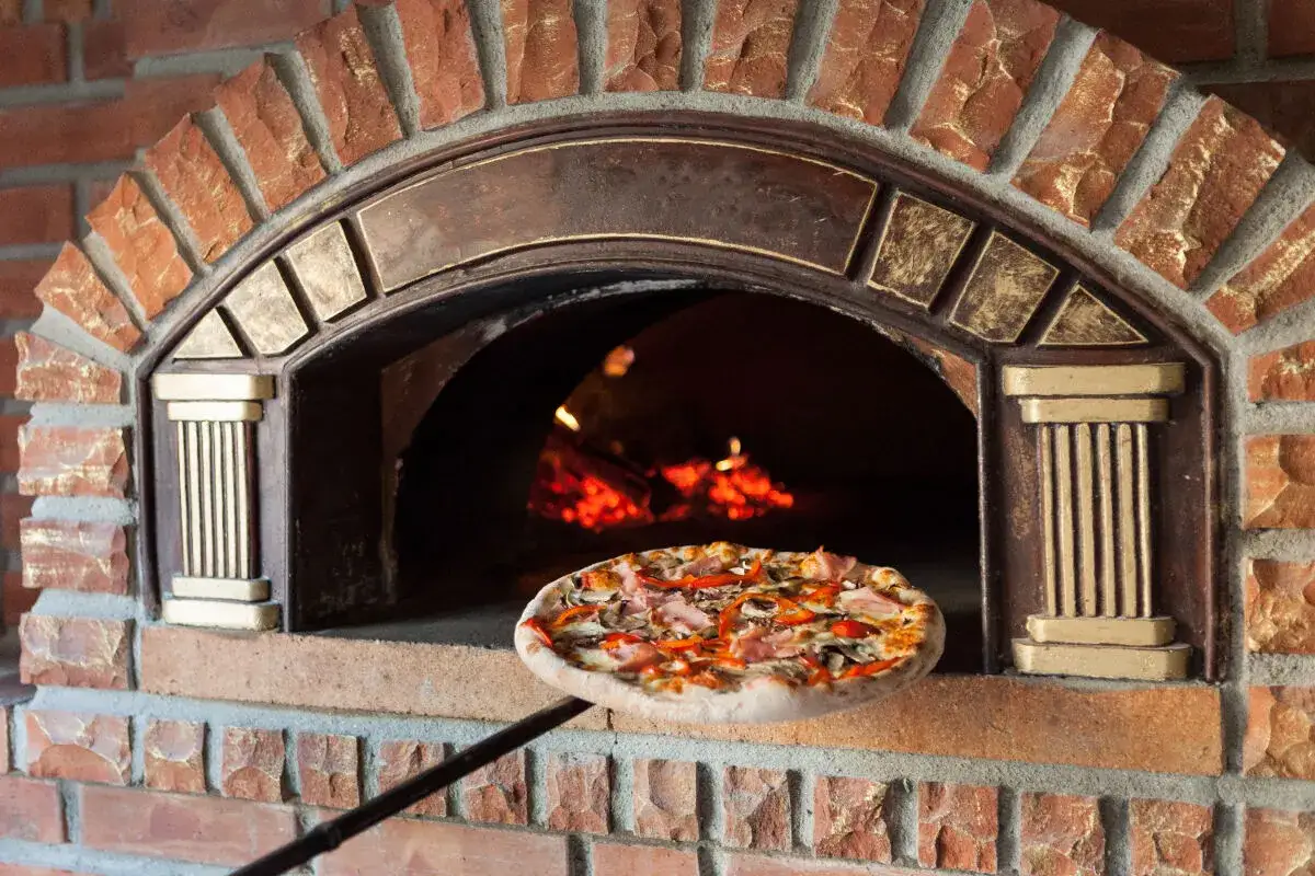 Une pizza garnie est sur le point d'entrer dans un four à pizza extérieur en briques rouges. Le feu crépite à l'intérieur.
