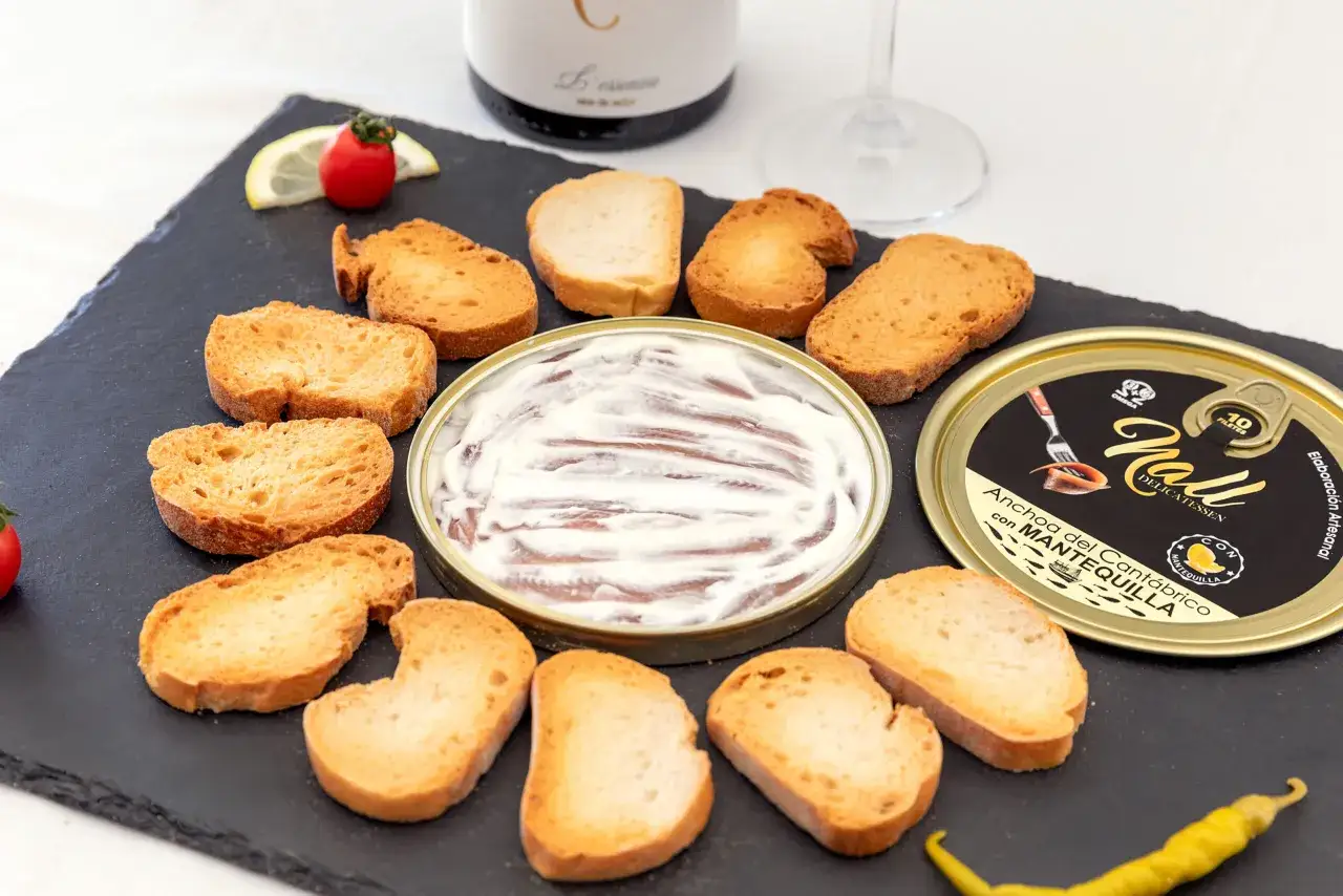 Una lata abierta de las mejores anchoas del mundo con mantequilla, rodeada de tostas, un lim&oacute;n, un tomate cherry y un pimiento.