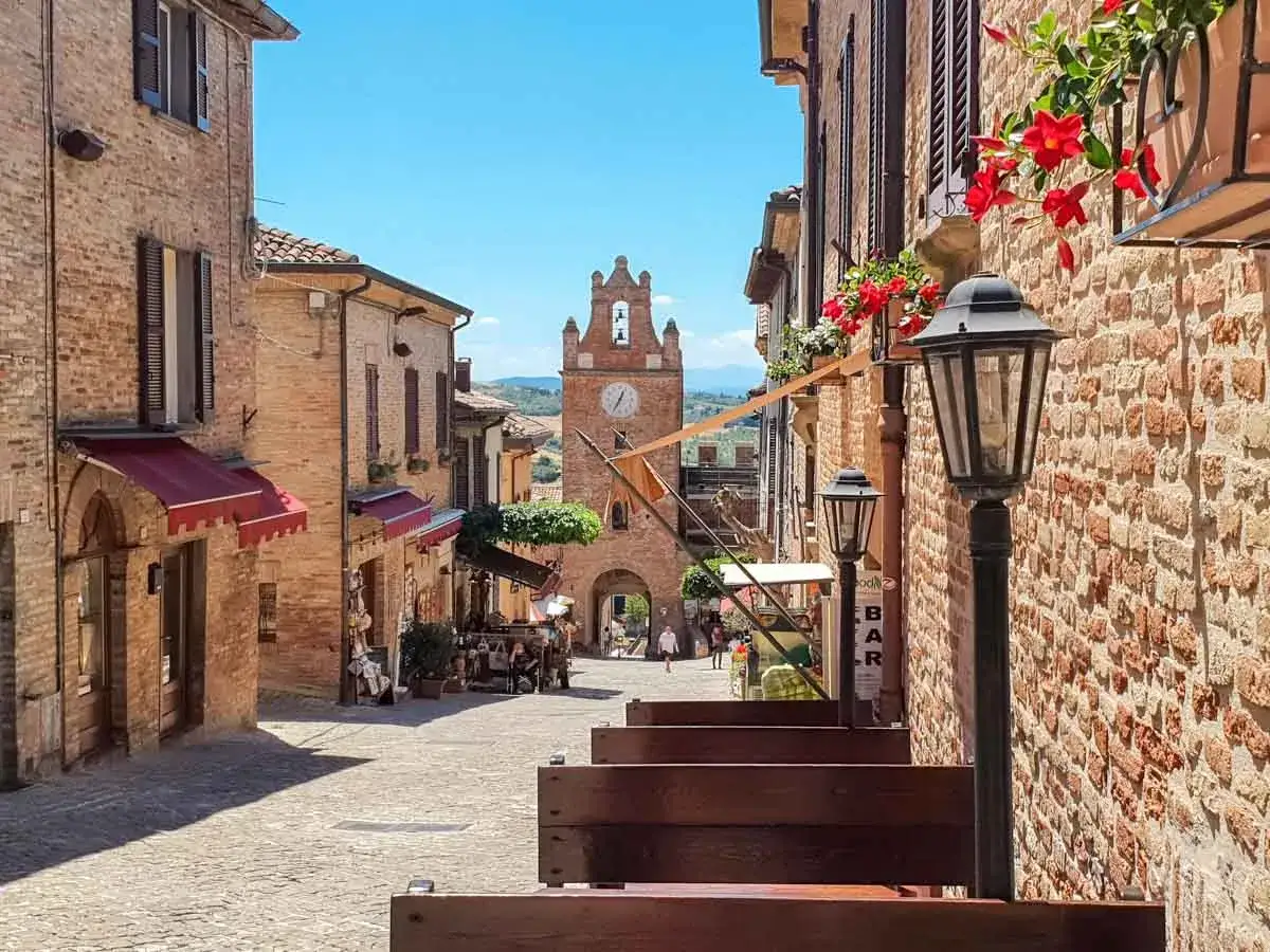 Un vicolo pittoresco a Gradara, Marche, con edifici in mattoni, fiori rossi e una torre dell'orologio in lontananza.