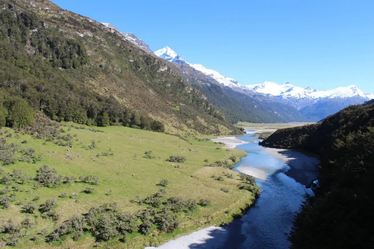 Un paysage de montagne, digne d'une peinture, avec une rivi&egrave;re aux eaux bleues serpentant &agrave; travers une vall&eacute;e verdoyante, sous un ciel clair.