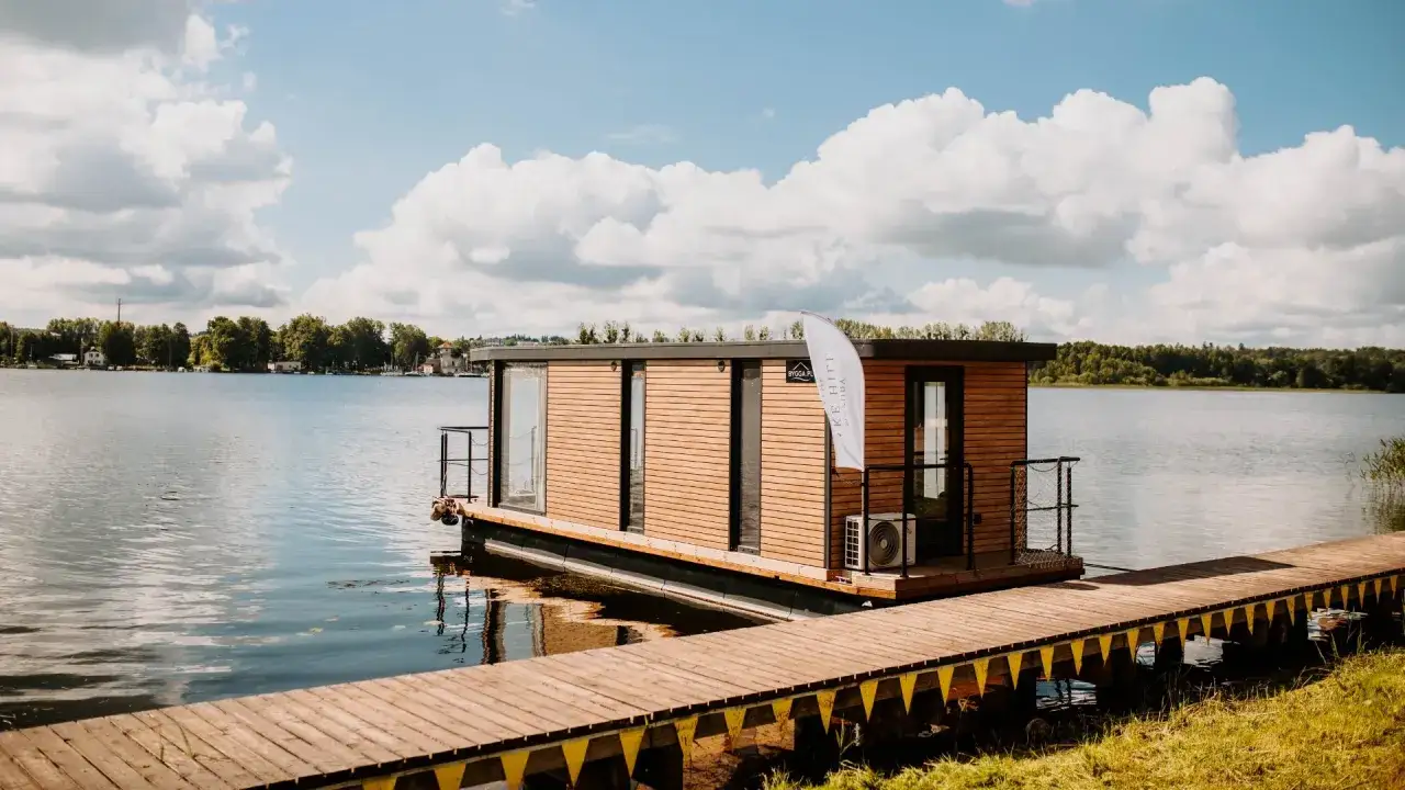 Idealne miejsce na mazury na weekend: nowoczesny dom na wodzie z drewnianym pomostem, otoczony jeziorem i zielenią.