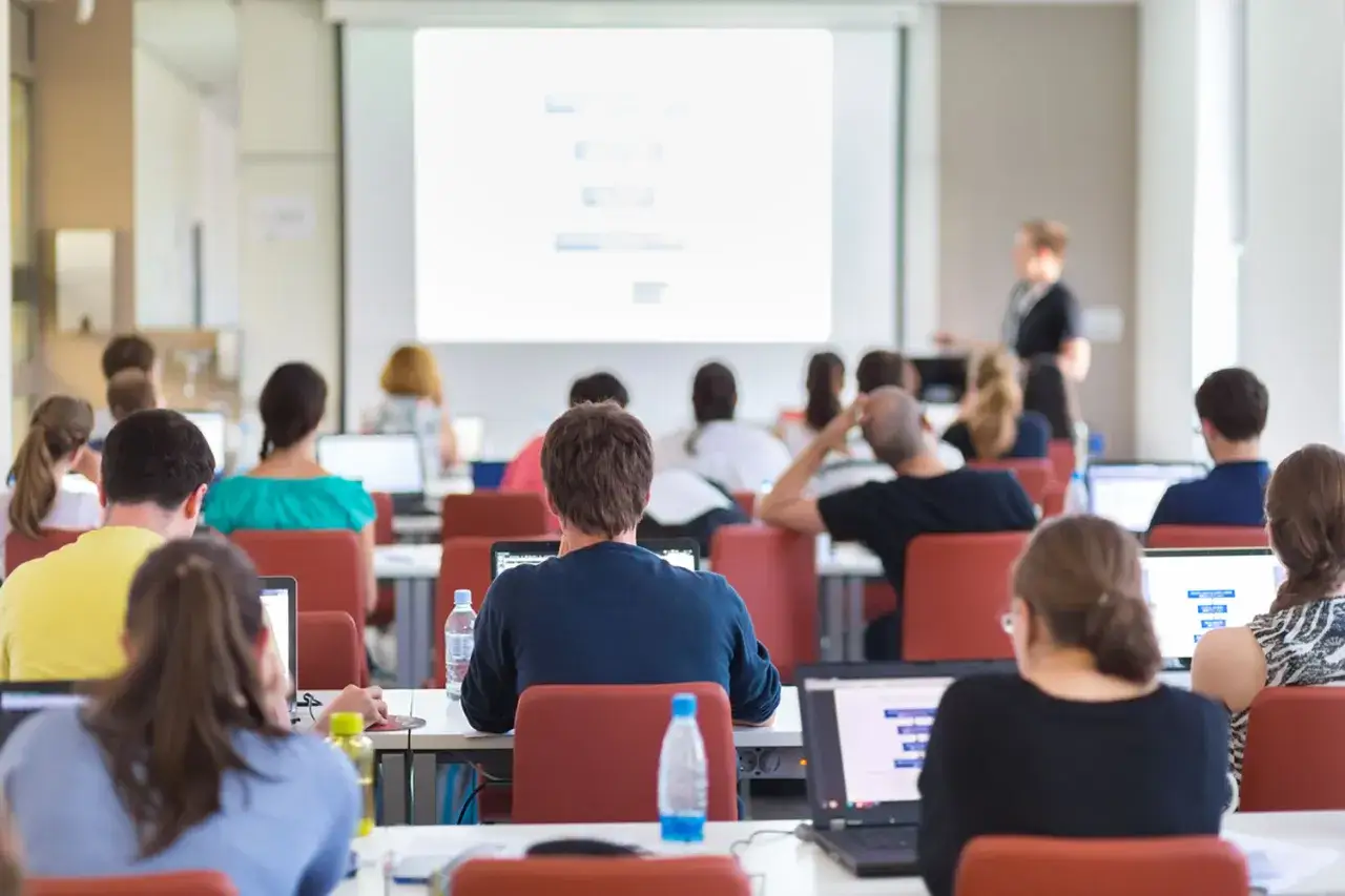 Asistentes en una sala de **formaci&oacute;n presencial**, atentos a la presentaci&oacute;n en pantalla y sus ordenadores port&aacute;tiles.