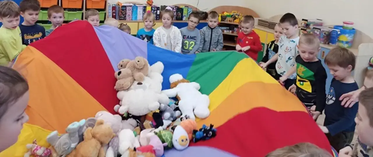 Dzieci świętują dzień misia, bawiąc się pluszakami pod kolorowym spadochronem.