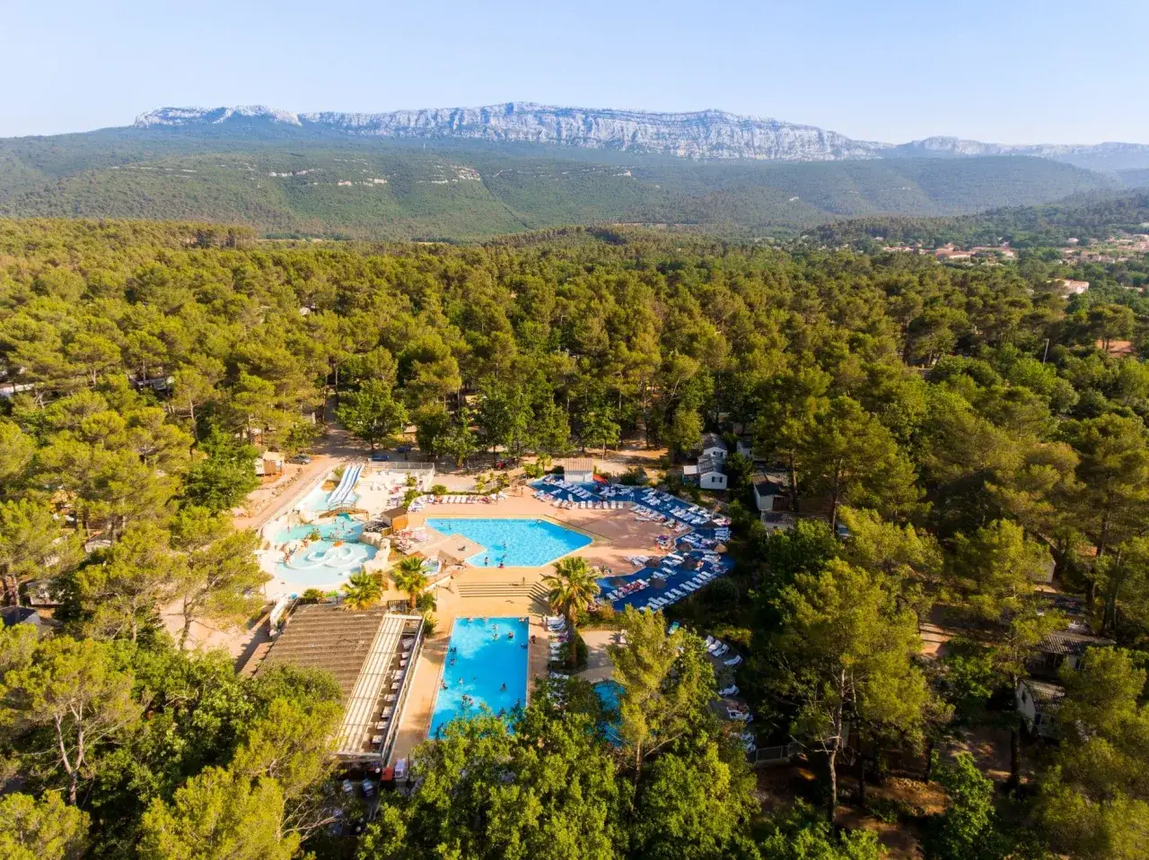 Aerial view of a resort with pools and slides, nestled among trees with a mountain range in the background, near Saint-Maximin-la-Sainte-Baume.