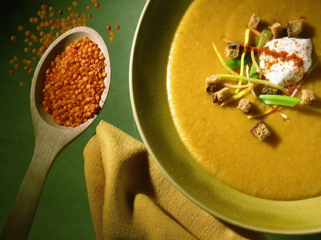 Eine cremige Linsensuppe, garniert mit Croutons und einem Klecks Sauerrahm. Daneben ein Holzl&ouml;ffel voller roter Linsen, bereit f&uuml;r k&ouml;stliche Linsen Rezepte.
