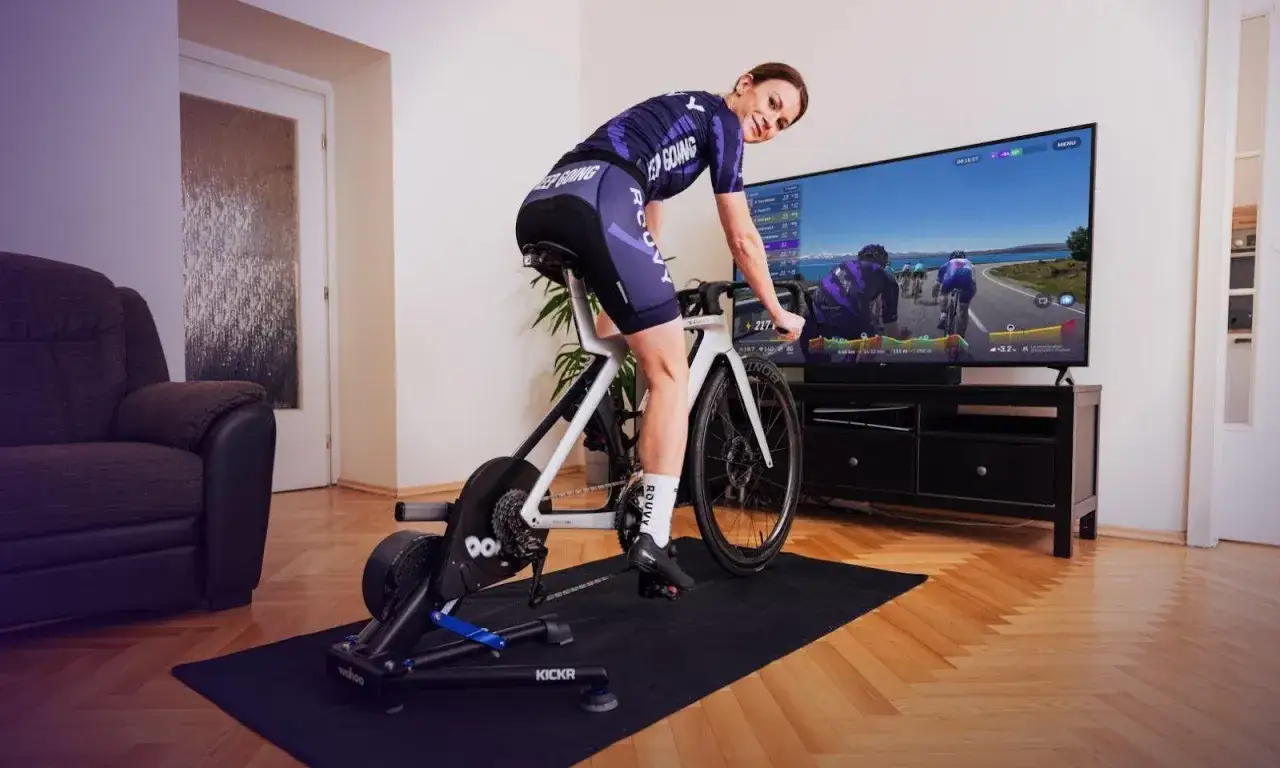 Kobieta wykonuje ćwiczenia aerobowe na rowerze stacjonarnym, śledząc wirtualny wyścig na ekranie telewizora.