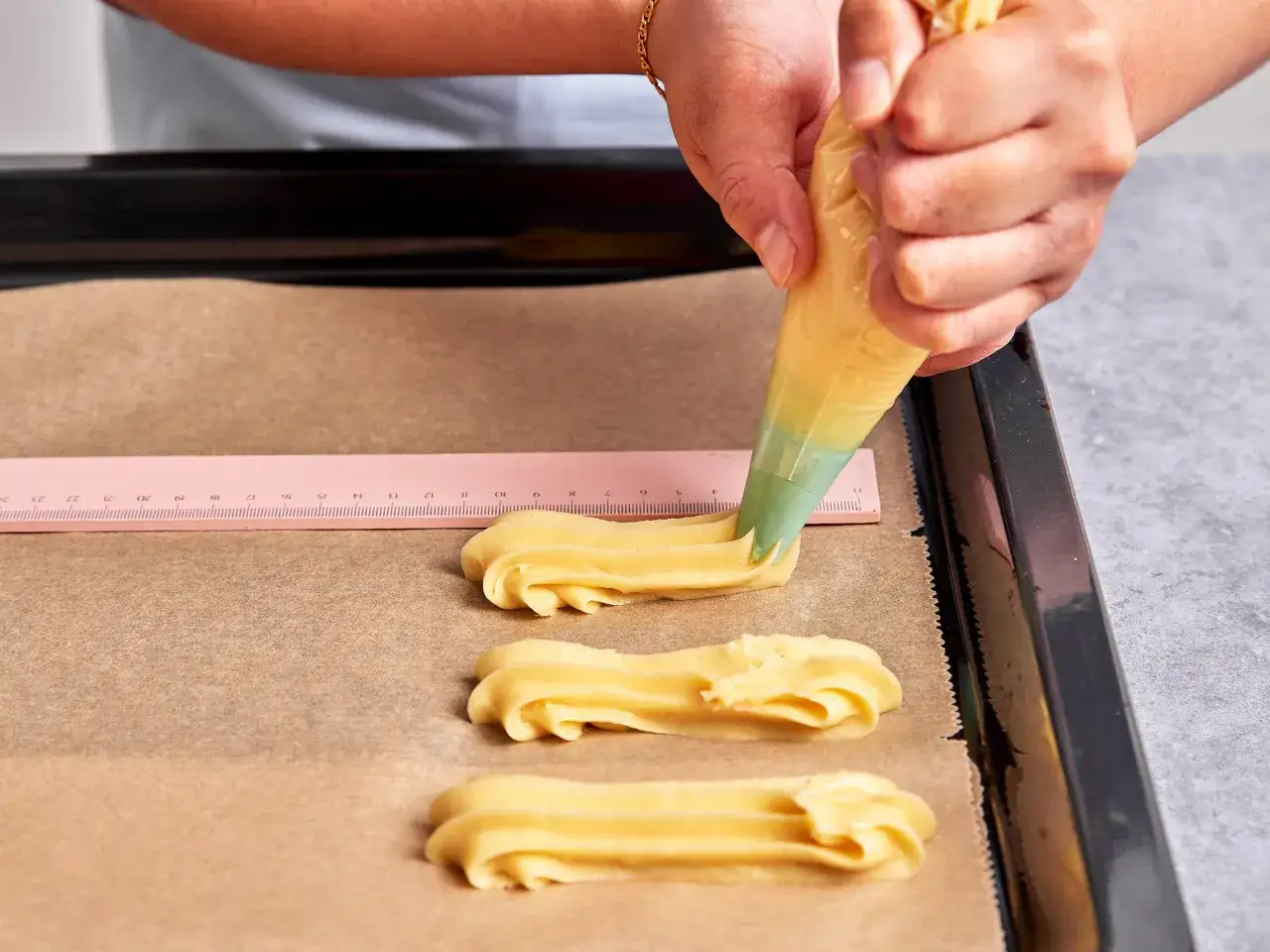 Mit einem Spritzbeutel werden Teiglinge f&uuml;r das Eclair Rezept auf Backpapier gespritzt. Eine Lineal hilft bei der gleichm&auml;&szlig;igen Gr&ouml;&szlig;e.