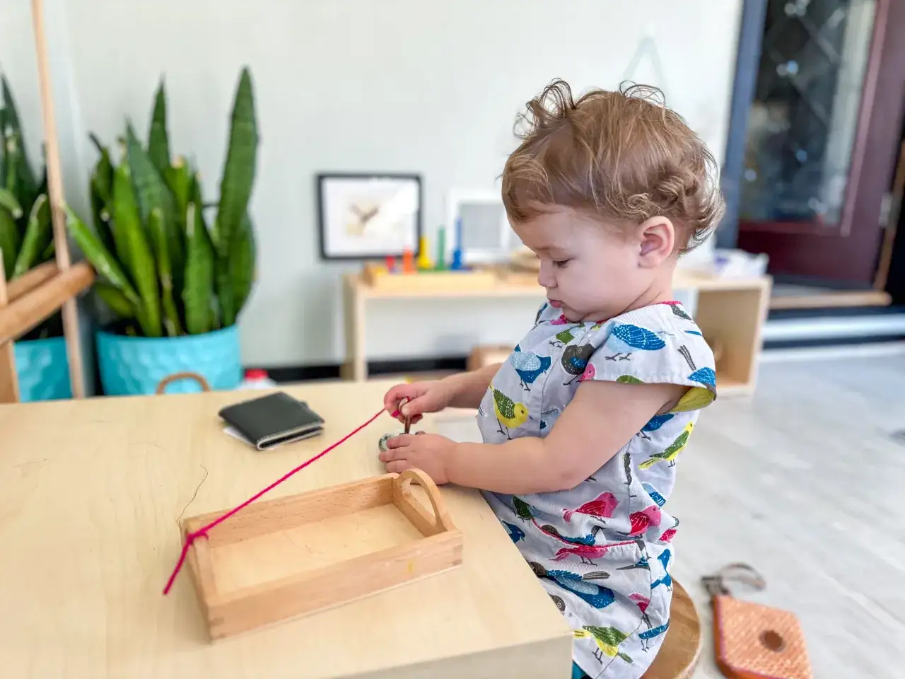 A toddler engages in Montessori baby activities, carefully threading a pink string through a small wooden object.