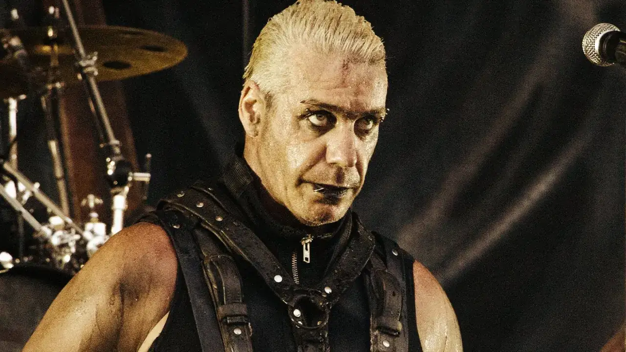 Till Lindemann młody, spocony, z kolczykami w twarzy, w skórzanym gorsecie, na tle perkusji.