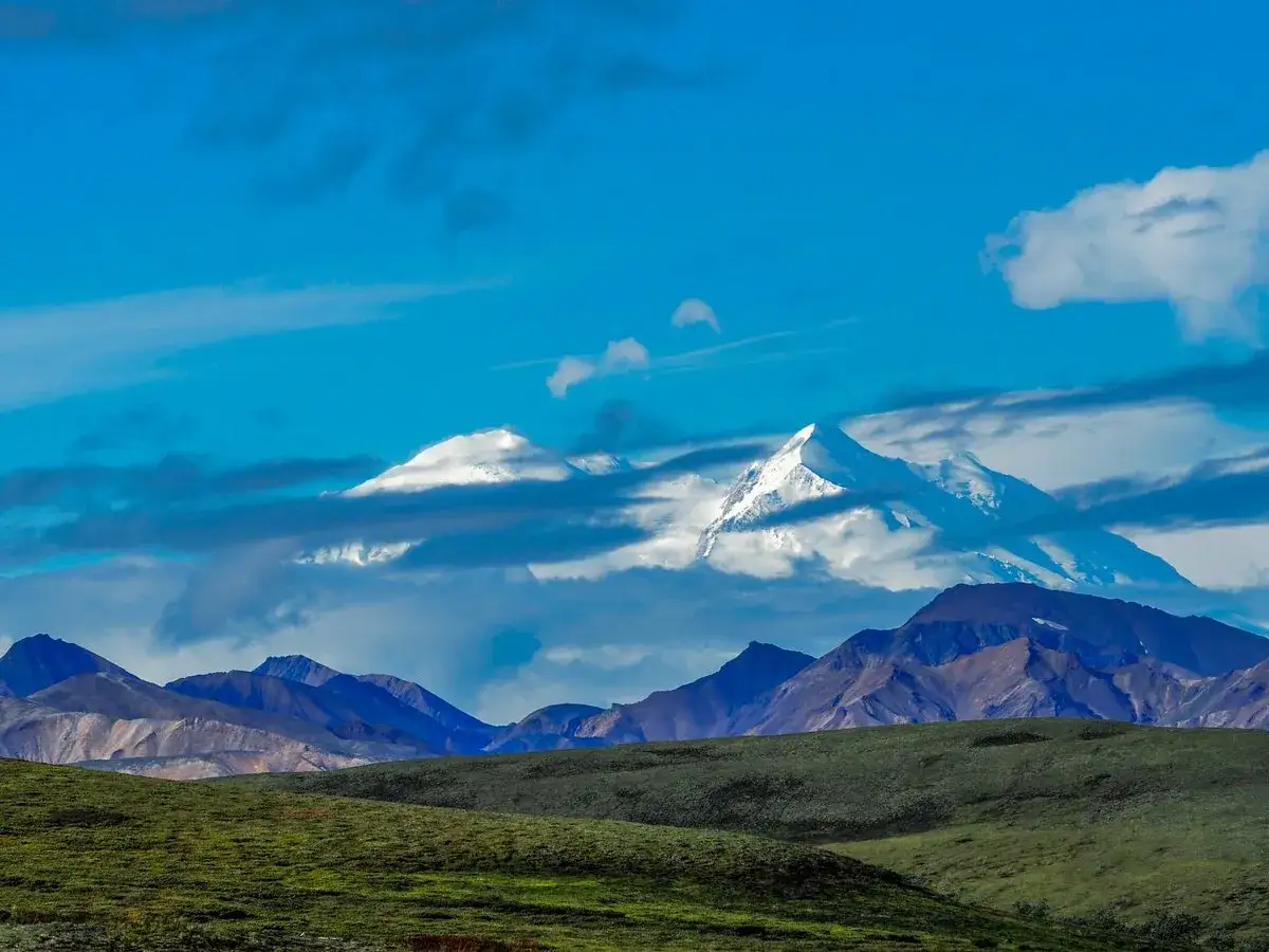 Denali, najwyższy szczyt Ameryki P&oacute;łnocnej, wyłania się z chmur nad zielonymi wzg&oacute;rzami.