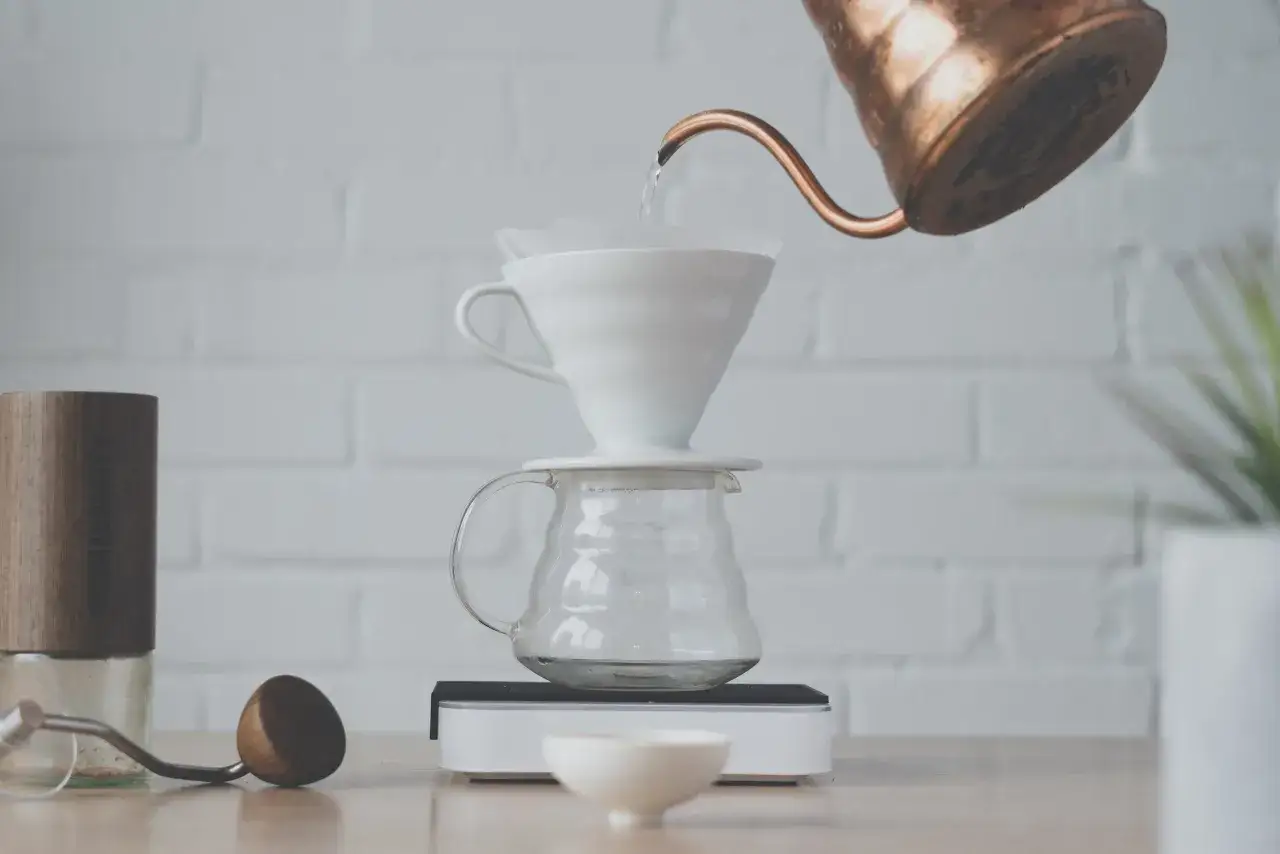 Vatten h&auml;lls fr&aring;n en kopparkanna &ouml;ver ett filter i en vit tratt, f&ouml;r att g&ouml;ra dropp kaffe. En kaffekvarn och en skopa syns bredvid.
