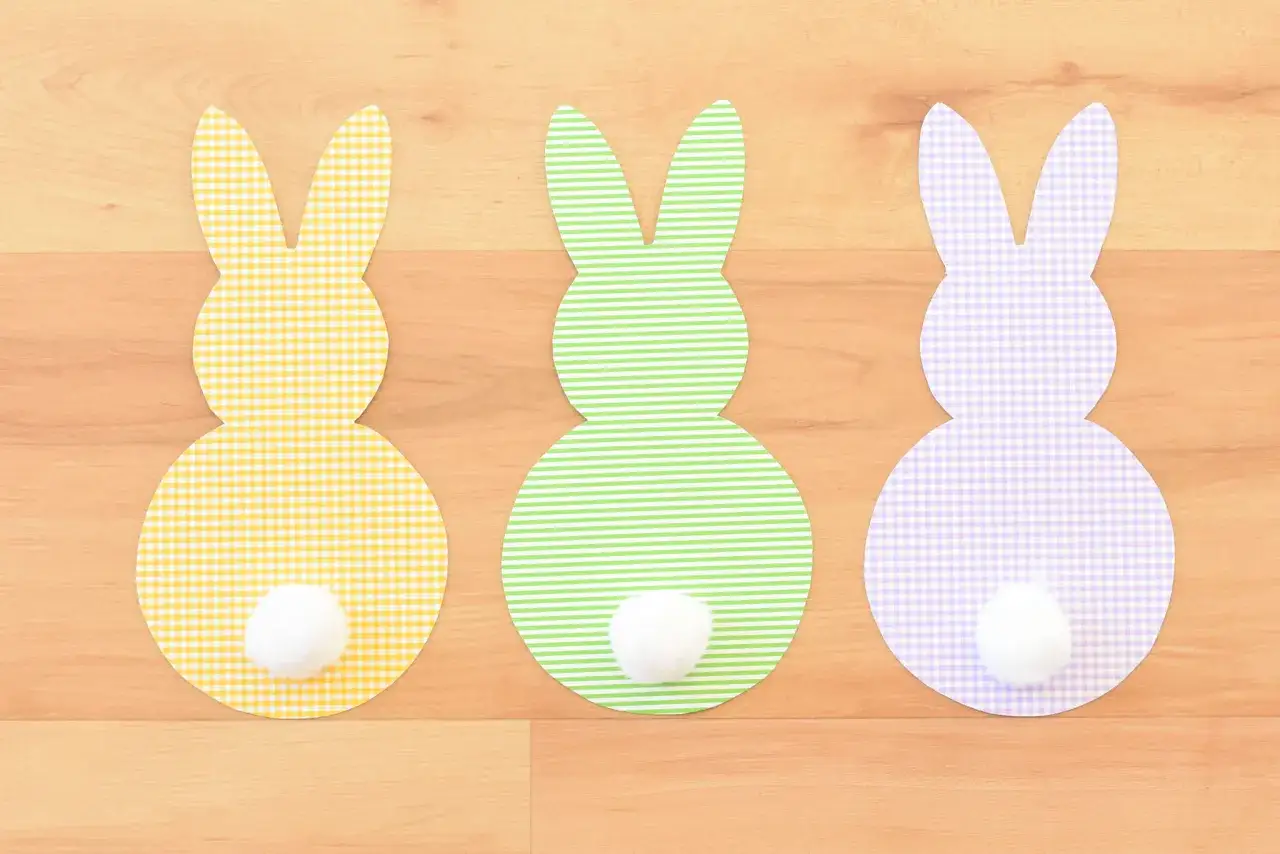 Drei bunte Papierhasen mit flauschigen Schw&auml;nzen stehen nebeneinander auf Holz. Ein gelber, ein gr&uuml;ner und ein lila hase.
