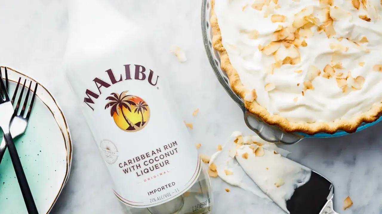 Butelka Malibu, karaibski rum z likierem kokosowym, obok pysznego ciasta z kremem i wiórkami kokosowymi. Idealny alkohol kokosowy na deser.