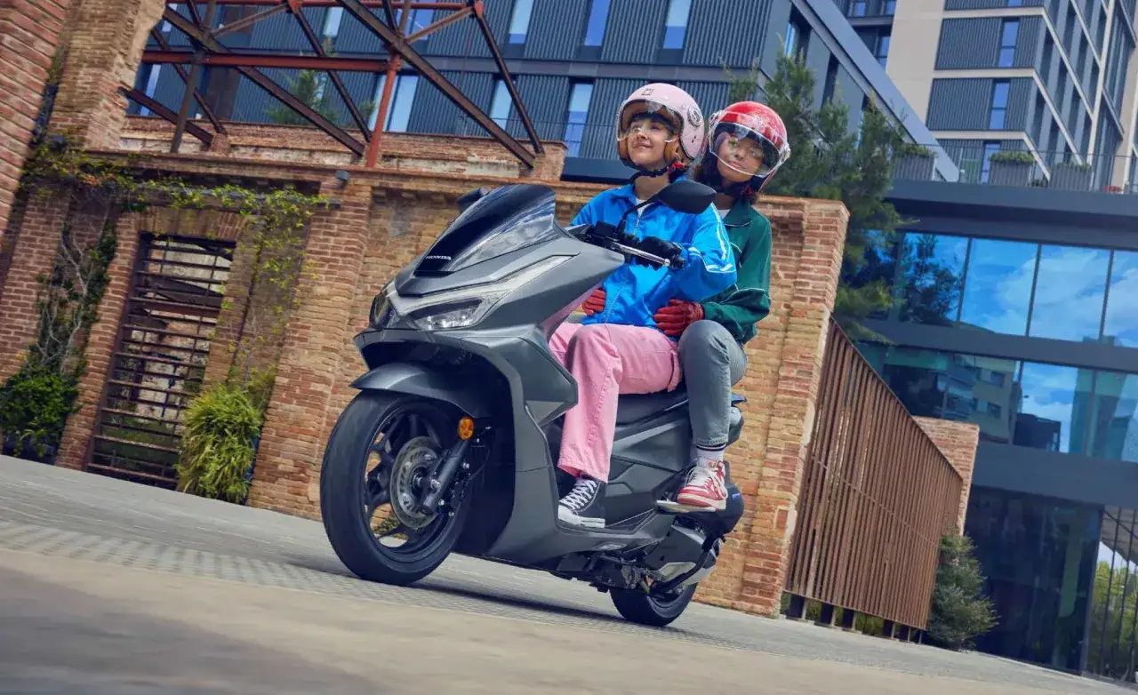 Dos jóvenes disfrutan de un paseo en una a1 moto, con cascos rosa y rojo, frente a un edificio moderno y una estructura de ladrillo.