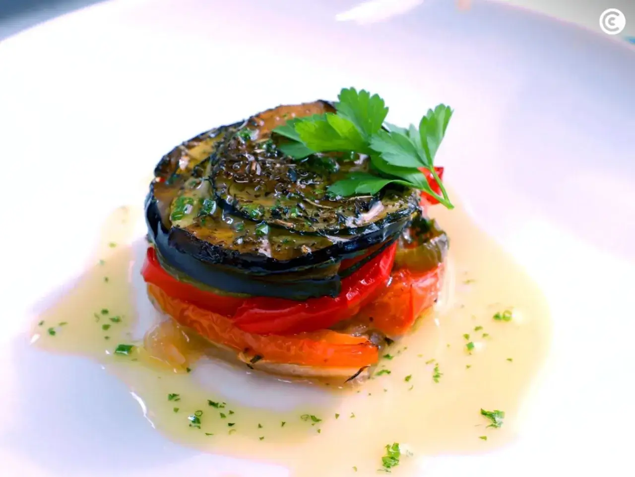 Delicioso ratatouille apilado, mostrando los ingredientes frescos: berenjena, tomate y pimiento, ba&ntilde;ado en una salsa dorada y adornado con perejil.