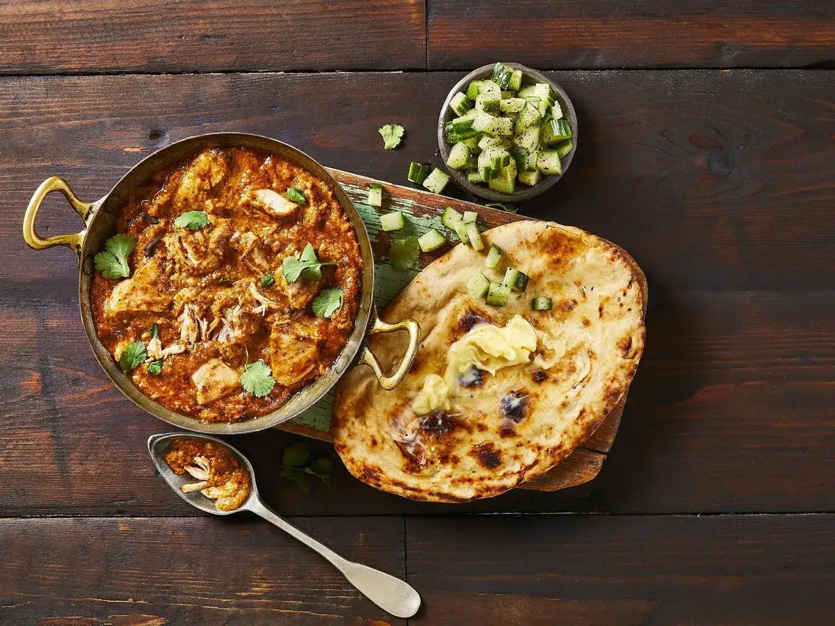 En sk&aring;l med kr&auml;mig butter chicken, serverad med naanbr&ouml;d, gurksallad och en sked full av smak.