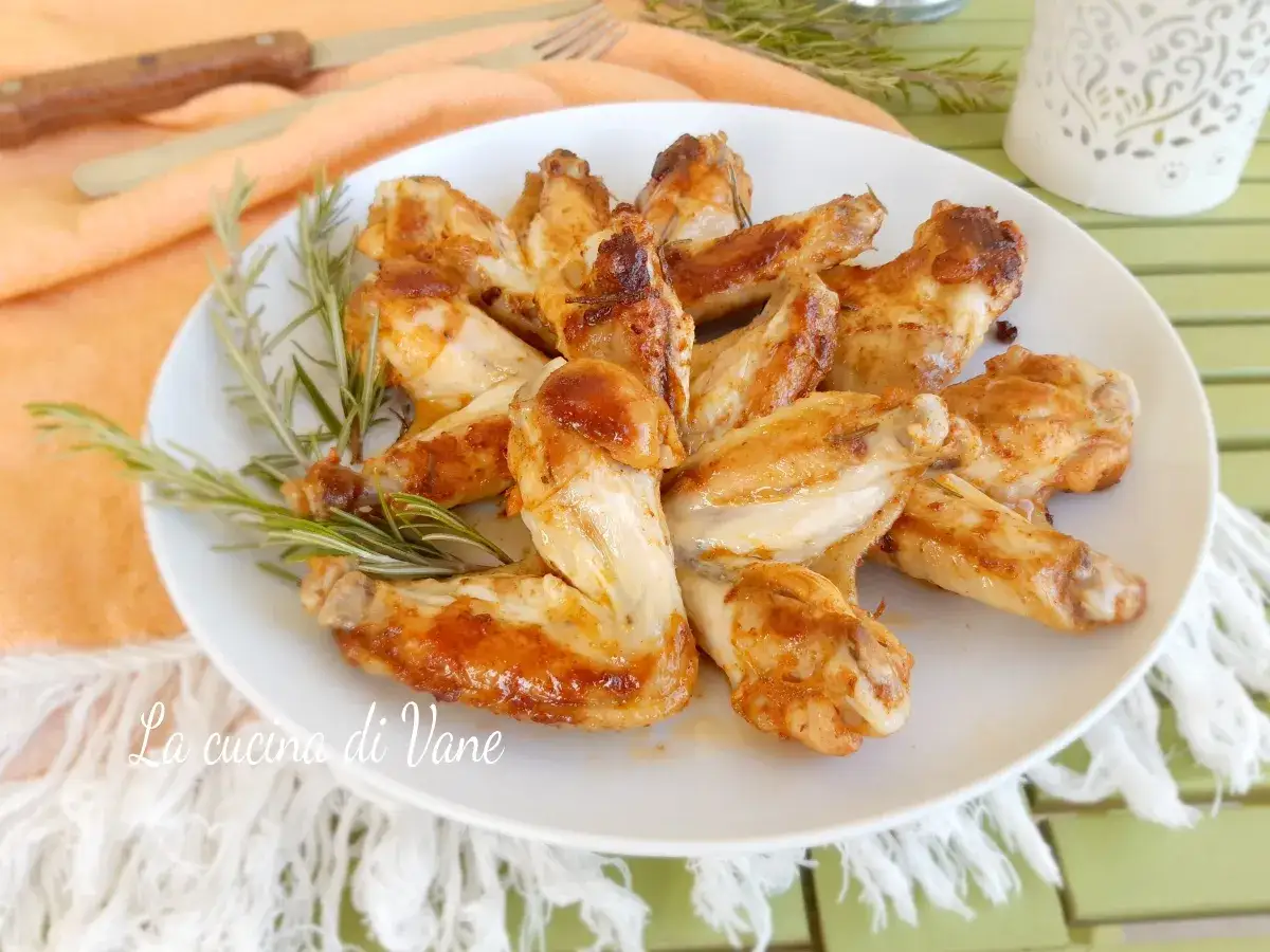 Alette di pollo in padella, dorate e succulente, guarnite con rosmarino fresco su un piatto bianco.