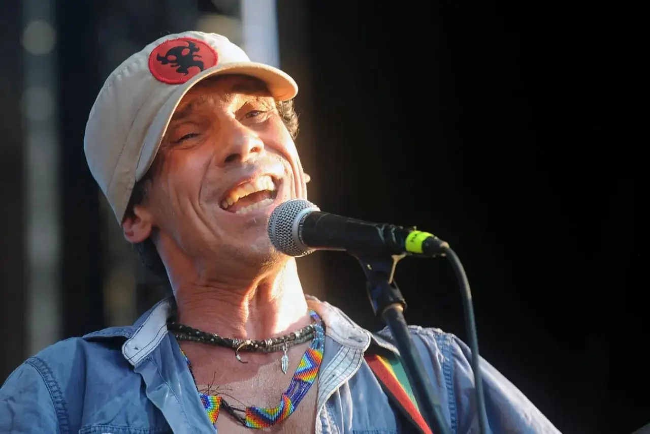 Un cantante spagnolo attuale con un cappellino e collane colorate canta con passione al microfono.
