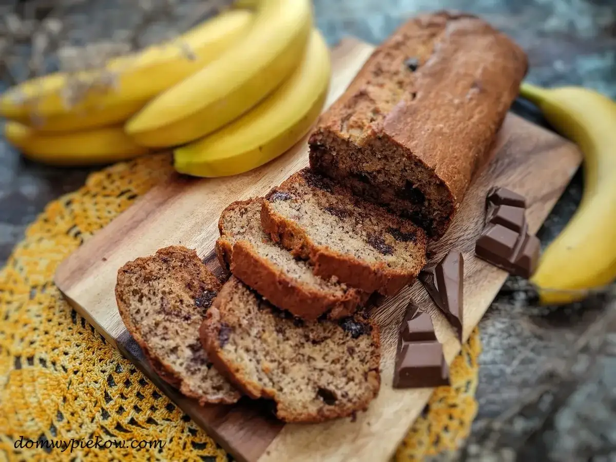 Pyszny chlebek bananowy z czekoladą, pokrojony w plastry, obok świeżych bananów i kostek czekolady.