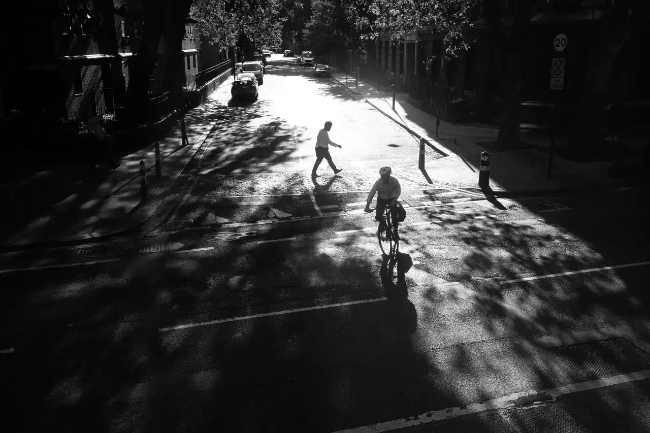 A cyclist and pedestrian cross a sun-dappled street, captured with a Voigtlander Color-Skopar 35mm lens, creating dramatic shadows.