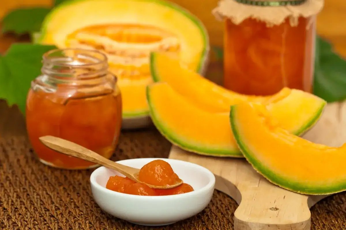 Marmellata di melone in vasetti e ciotolina con cucchiaino, accanto a fette di melone fresco.