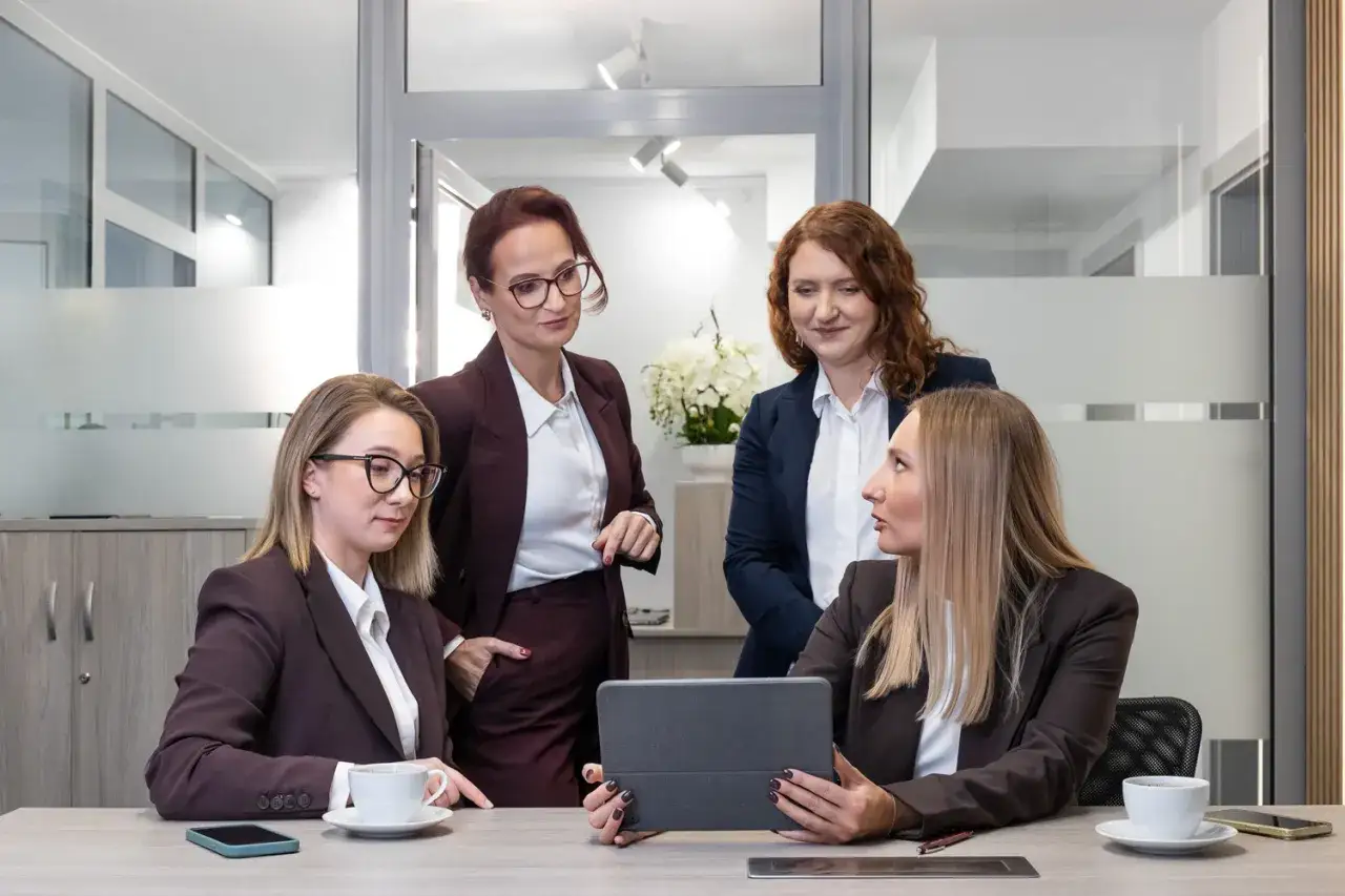 Cztery kobiety w garniturach dyskutują przy biurku z tabletem i kawą.