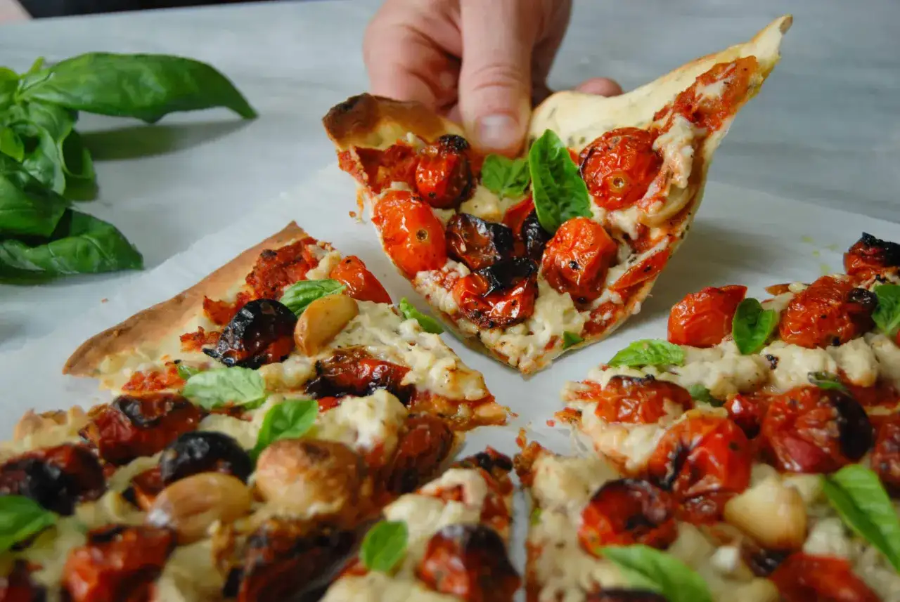 Eine Hand hebt ein St&uuml;ck Pizza mit Kirschtomaten, Knoblauch und Basilikum. Der vegane Pizza Belag sieht k&ouml;stlich aus.