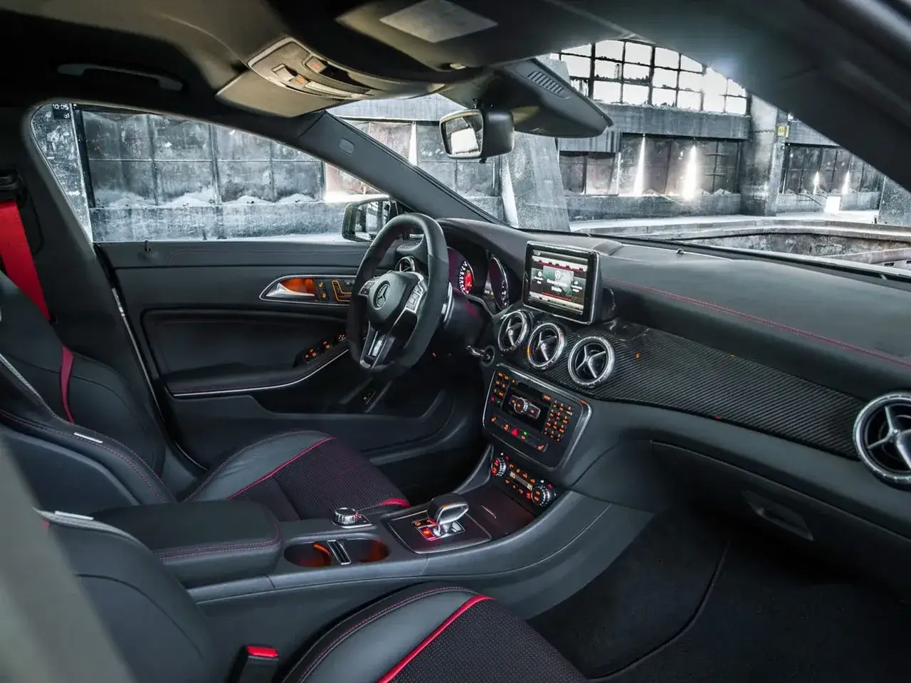 Wnętrze sportowego Mercedesa CLA 45 AMG z czarną tapicerką i czerwonymi przeszyciami. Kierownica, deska rozdzielcza i konsola środkowa.