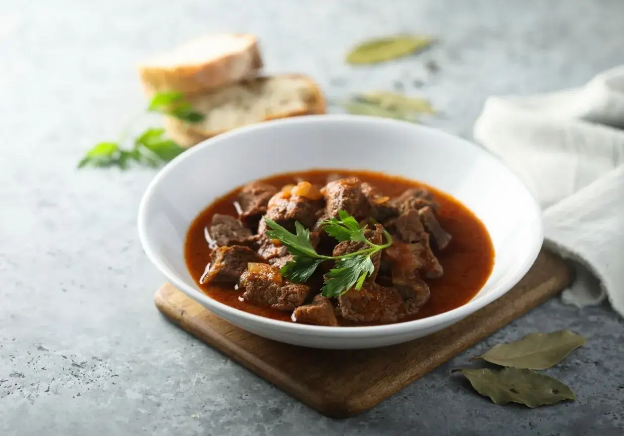 Aromaticzny, tradycyjny gulasz wieprzowy w białej misce, udekorowany natką pietruszki. Obok kromki chleba i liście laurowe.