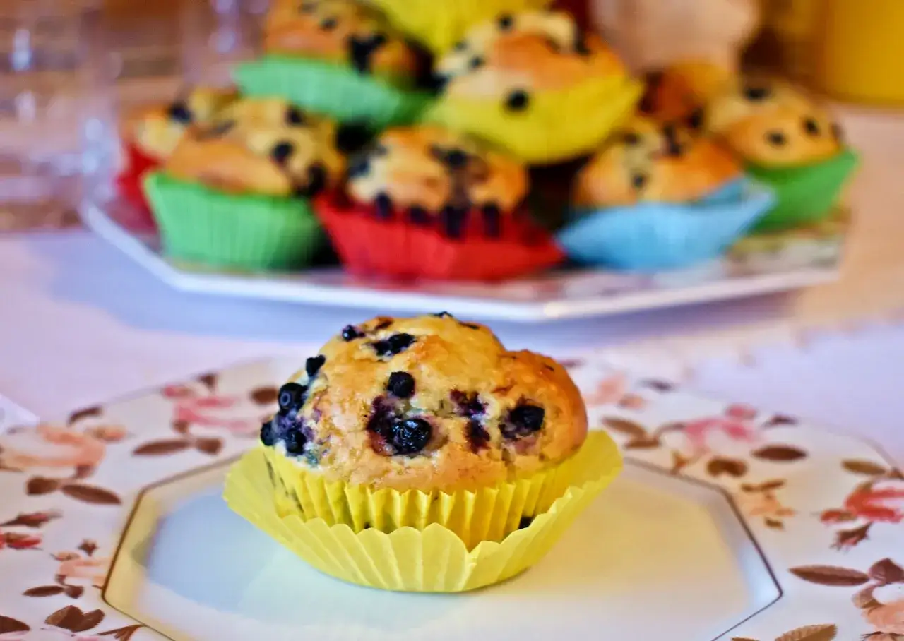 Przepis na wilgotne i puszyste muffinki jagodowe. Na pierwszym planie jeden muffinek w żółtej papilotce, w tle stos kolorowych muffinek.