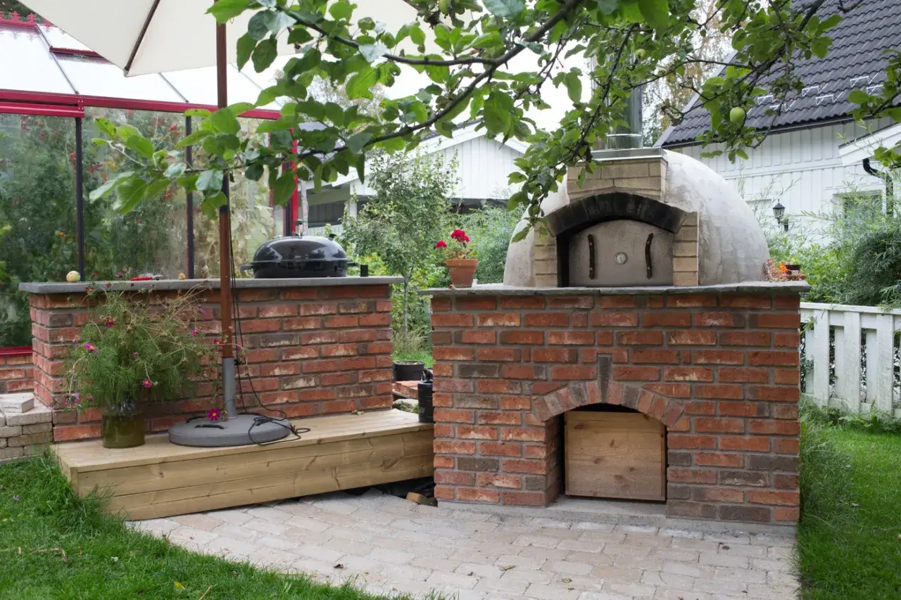 En vacker mura bakugn i trädgården, redo för pizza. Bredvid står en grill och ett växthus.