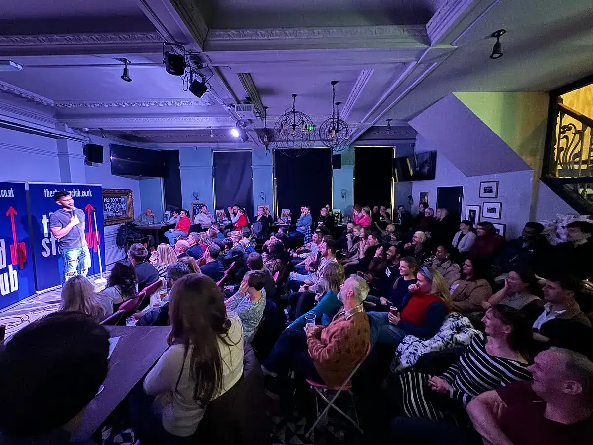 En brittisk stand up komiker framför en publik i en mörk lokal.