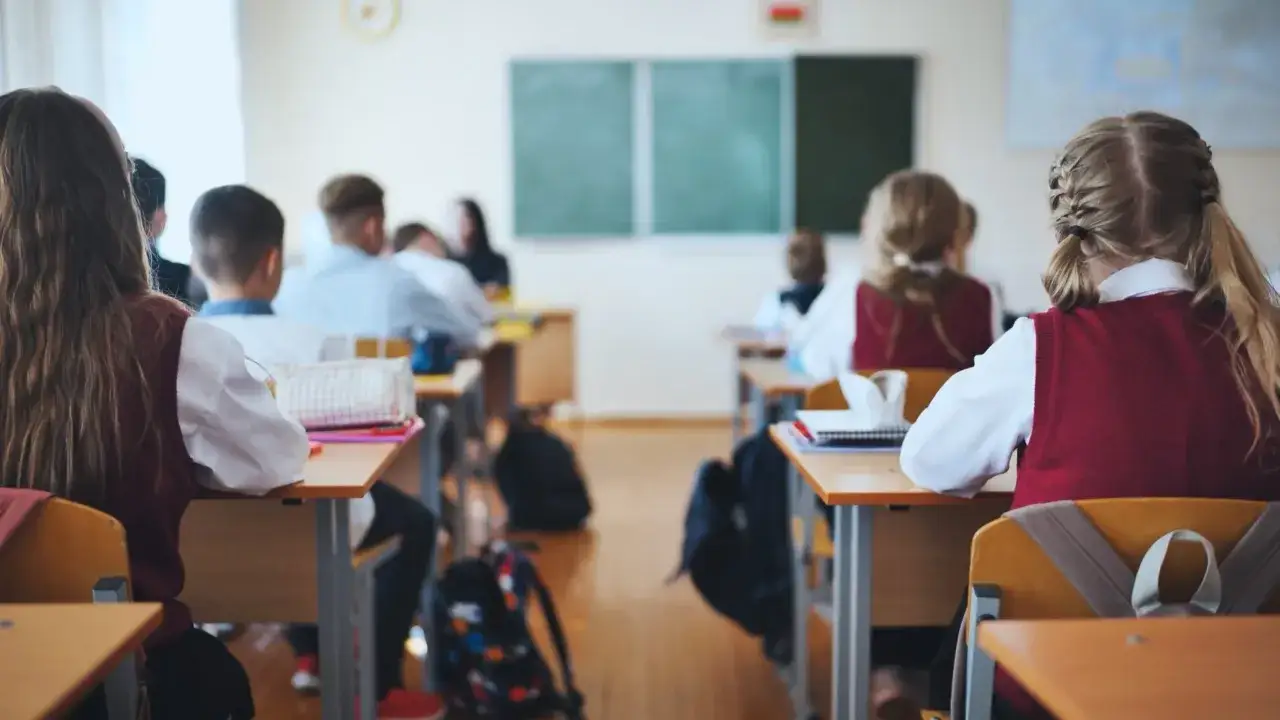 Uczniowie siedzą w ławkach, przygotowując się do lekcji. W tle widać tablicę. Trudno określić, ile jest klas w liceum, ale sala lekcyjna jest pełna.
