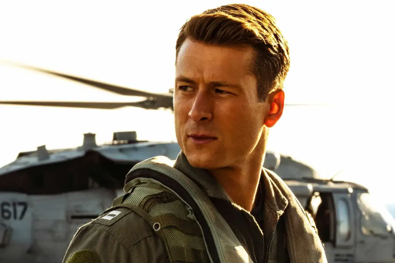 Młody pilot w mundurze, gotowy na misję w stylu Top Gun 3, z helikopterem w tle.