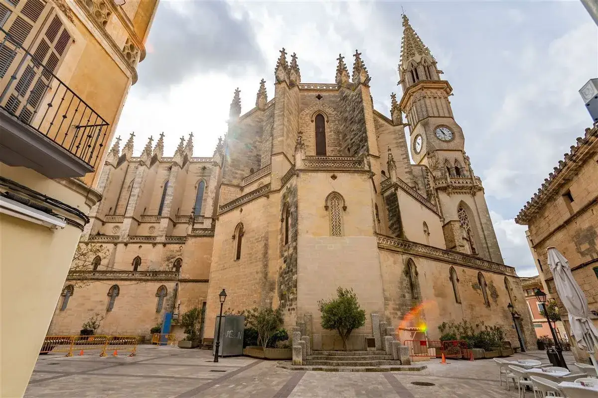 Imposante Kirche mit Turmuhr im Herzen der **Manacor Altstadt**. Sonnenschein bricht durch Wolken.