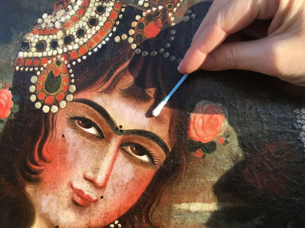Main d'un restaurateur nettoyant délicatement un tableau ancien avec un coton-tige, révélant la beauté d'un visage orné.