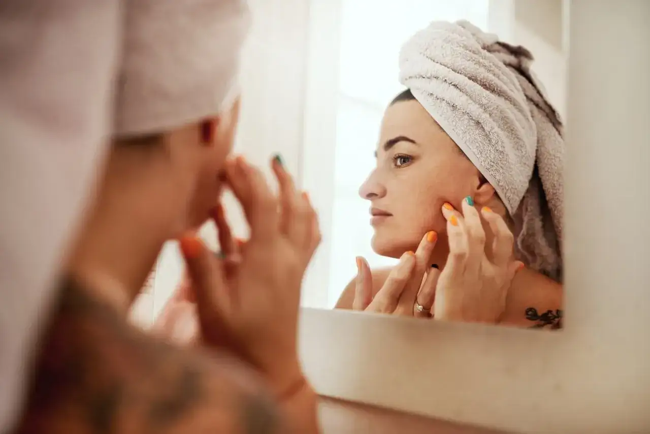 Mujer con toalla en la cabeza se mira al espejo, tocando su rostro. Aha, ¡qué es esa pequeña imperfección!