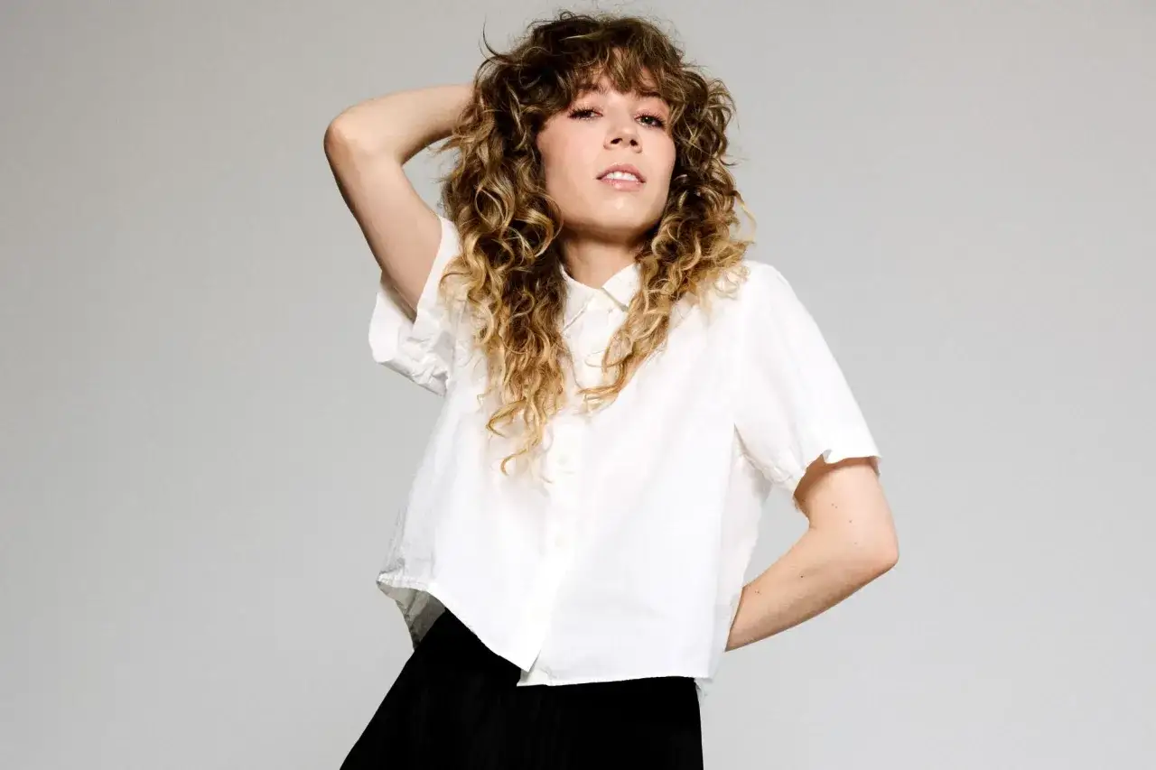 Jennette McCurdy con capelli ricci biondi, indossa una camicia bianca e una gonna nera, con una mano tra i capelli.