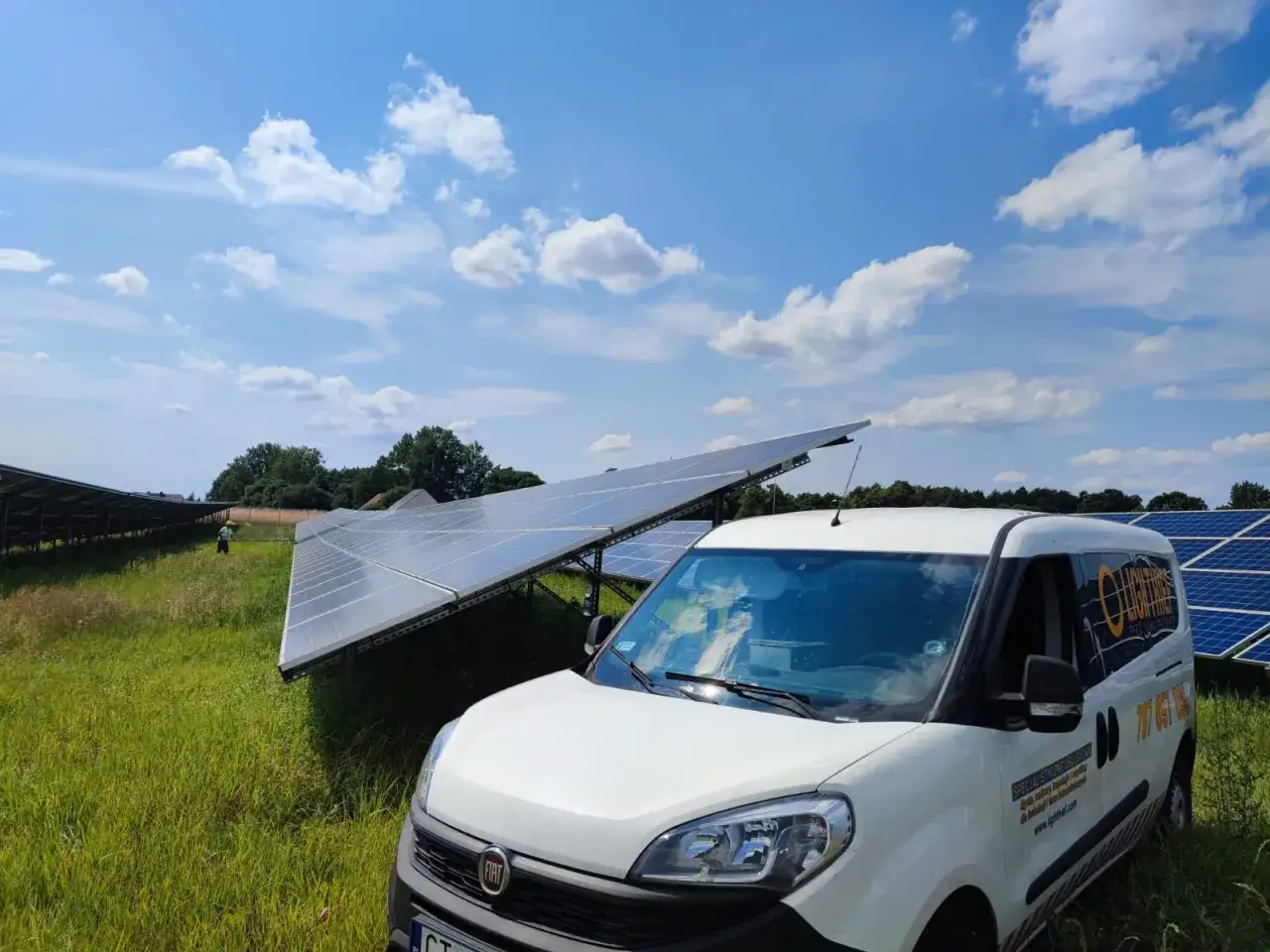 Biały bus serwisowy obok paneli fotowoltaicznych. Sprawdź, jaka jest serwis fotowoltaiki cena i zadbaj o swoją instalację.