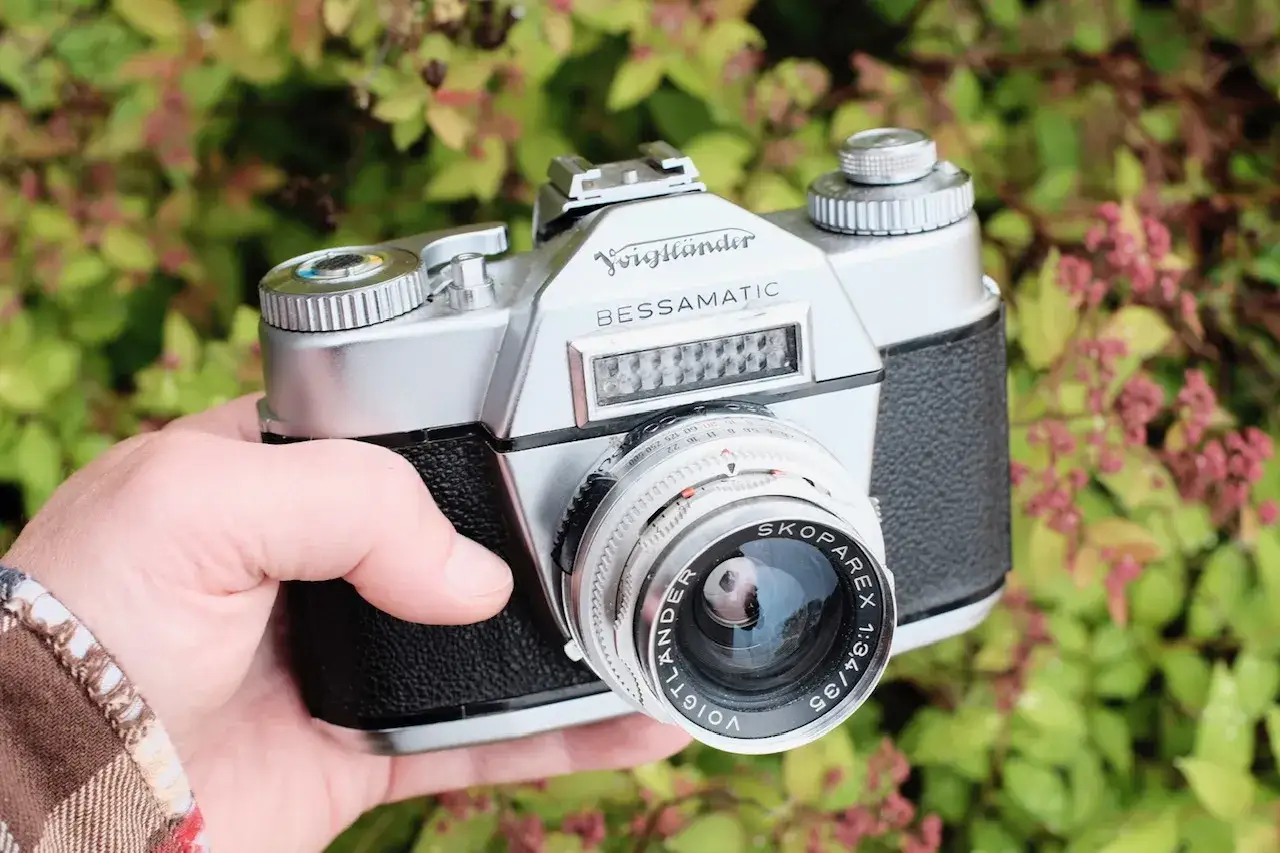 A hand holds a vintage silver Voigtlaender Bessamatic camera with a Skoparex lens, set against a backdrop of lush green foliage.